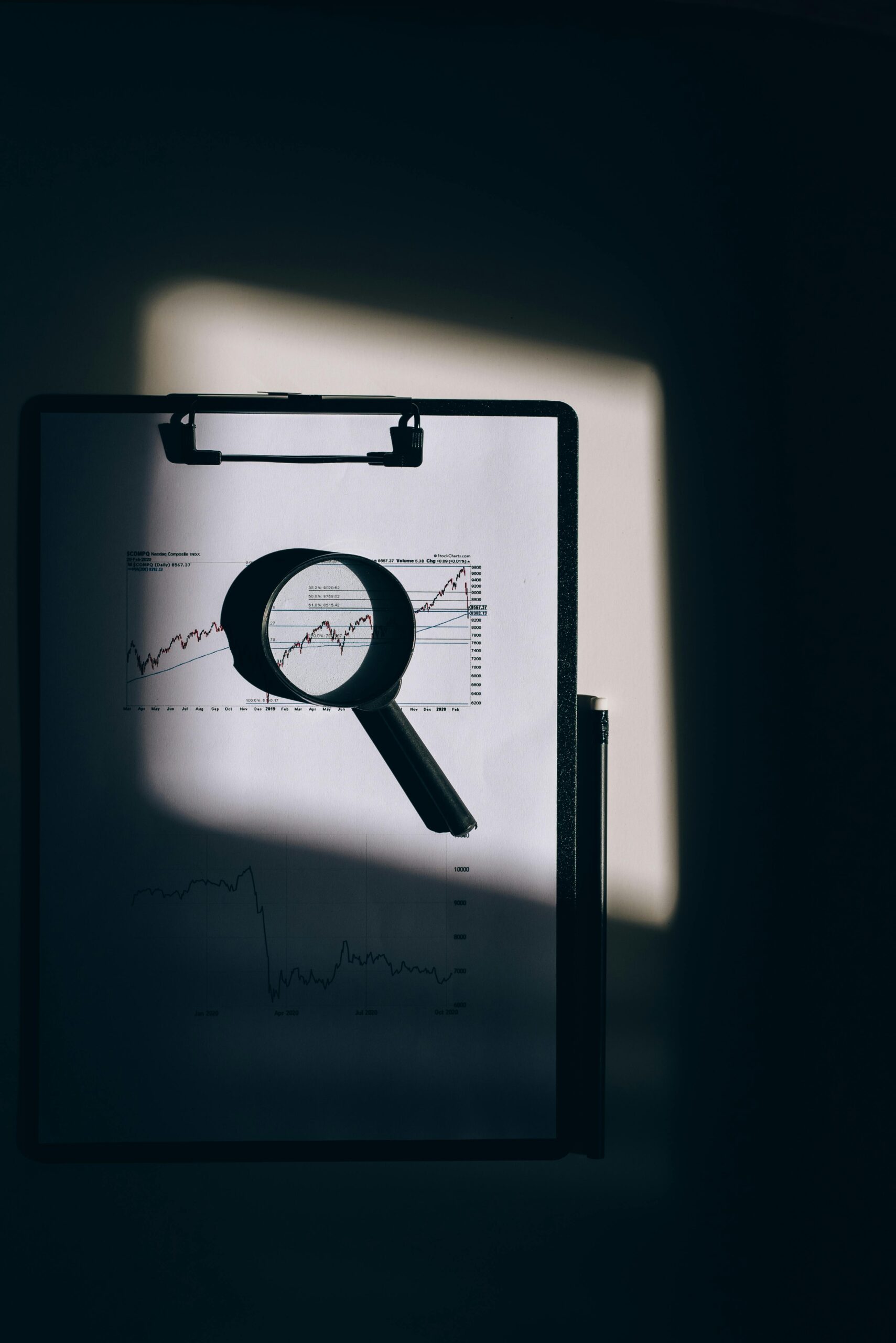 A magnifying glass lays over a financial chart on a clipboard, casting intriguing shadows.